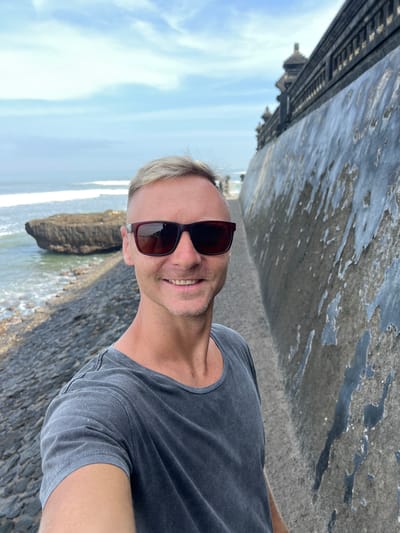 A man with short, blonde hair and sunglasses takes a selfie on a cobblestone path with the ocean on his left and a textured, weathered wall on his right. The wall appears to be part of a large, dark, ornate structure, possibly a temple or historical building, with decorative elements visible at the top. The sky is overcast with a few streaks of blue. In the background, there's a large rock formation in the ocean, and waves are breaking on the shore. The man is wearing a dark grey t-shirt. The location is likely the Tanah Lot temple complex in North Kuta, Bali, Indonesia, known for its coastal setting and distinctive architecture.