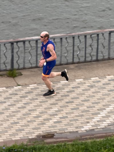 A man in a blue and black athletic outfit with orange trim is captured in motion, leaning forward as if running or mid-stride. He has a shaved head and appears to be wearing dark sunglasses. His left leg is extended behind him, and his right leg is bent at the knee. He is wearing black running shoes with white soles. The man is positioned on a paved walkway made of rectangular bricks in varying shades of grey and beige. To his left, there is a body of water, possibly a lake or river, with a dark, wavy surface. Adjacent to the walkway, on the right side of the frame, is a patch of vibrant green grass with a few small blue flowers. A dark metal railing with horizontal bars runs along the edge of the water. The lighting suggests it is daytime. The scene is set outdoors, likely in a park or recreational area, possibly near a waterfront. The man's posture indicates he is engaged in physical activity.