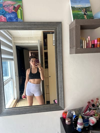 A reflection in a grey-framed mirror captures a young woman in workout attire—a black sports bra and white shorts—holding a cell phone in one hand and a red apple in the other. The mirror hangs on a white wall, positioned between a shelf filled with makeup items and a painting of whimsical figures. The reflection includes a glimpse of a bright, window-lit space with blinds, suggesting an interior. The reflected scene hints at a city location through the window, but there is no additional evidence indicating the location is Puebla, Mexico.