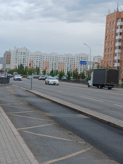 The image shows a multi-lane road in Astana, Kazakhstan, with several vehicles driving on it. In the foreground, there is a sidewalk and a section of the road with yellow diagonal markings, possibly indicating a no-parking zone. Several apartment buildings of varying architectural styles are visible in the background, some with beige facades and others with white facades and numerous windows. A large, modern orange-colored apartment building with many balconies is prominent on the right side of the image. Traffic lights, streetlights, and road signs are also present. The sky is overcast with clouds.