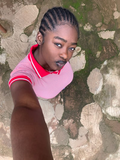 An overhead, high-angle selfie shows a young Black woman posing outdoors. She has braided hair and is wearing a red and white striped polo shirt. She is looking directly at the camera with a neutral expression. The background consists of cracked concrete with patches of moss. The scene appears to be located in a courtyard or patio area. The location in the city of Rafin Par, Nigeria is not specified in the image itself and cannot be determined from the visual information provided.