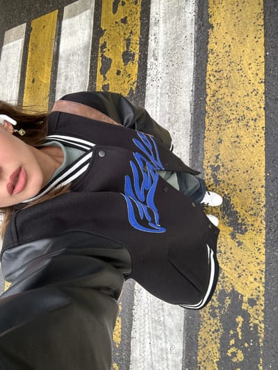 A person is standing on a crosswalk, indicated by the yellow and white stripes on the asphalt. They are wearing a black varsity jacket with blue flame-like embroidery on the front and white stripes on the collar. The jacket also features black leather sleeves. Underneath the jacket, a light green top is visible. The person is wearing white sneakers with black stripes. In their ears, white earbuds are visible. The lower part of their face is visible, showing pink lipstick and parted lips. The image appears to be taken from a high angle, looking down at the person. The lighting suggests it is daytime, and the asphalt appears wet, hinting at recent rain or damp conditions. The location is identified as Khimki, Russia.