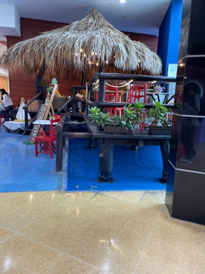 The image depicts a rustic outdoor seating area, possibly part of a restaurant or cafe, under a large thatched roof structure. The seating consists of red chairs around tables, with some tables positioned on a raised wooden platform. Potted plants are placed on the platform. In the background, there is a person with dark hair wearing a yellow top and another person with dark hair and a white top. A woman with long dark hair wearing a white shirt and dark pants is standing on the left side of the image, facing away from the camera. There are also decorative elements such as a statue or figurine on the left and a chalkboard menu. The floor is a mix of blue and tan tiles. The overall ambiance suggests a tropical or beach-themed setting. The location is identified as Porlamar, Venezuela.