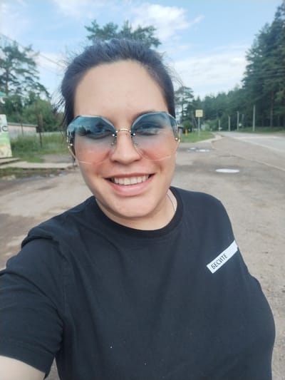 A medium shot captured from a slightly low angle shows a woman with dark hair pulled back, wearing large, rimless sunglasses with blue and pink gradient lenses. She is smiling broadly, revealing her teeth. The woman is wearing a black t-shirt with a white rectangular label on the left sleeve that reads "БЕСИТЕ" in black Cyrillic lettering. She is outdoors, with a paved road and a grassy verge visible in the background, lined by trees. The sky is partly cloudy. The location is identified as Rusalevka, Russia.