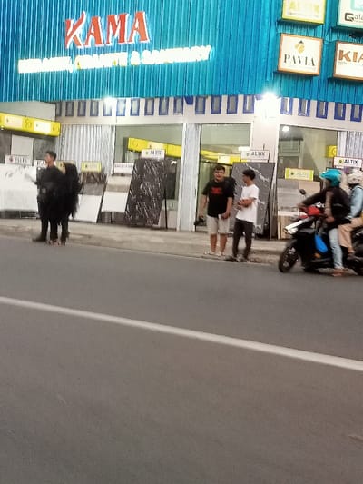 The scene is in front of a building in Pati, Indonesia, located at coordinates -6.753 latitude and 111.041 longitude. The building is a storefront with a blue and white facade. There is signage above the entrance for "KAMA Marmer, Granit & Saniteri". On the left side of the building there are four individuals walking away from the camera, two appear to be females wearing long black clothing. On the right side of the building, there is a motorcycle with three people riding. Two are wearing helmets, one with a blue helmet and the other with a black helmet. The motorcycle is traveling away from the camera.  A white painted line is in the center of the asphalt road. The scene is taken in the daytime.