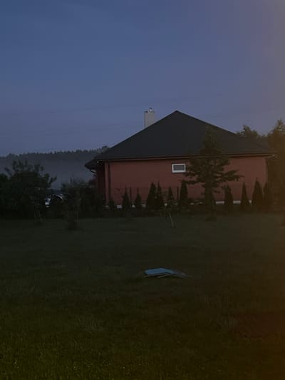 In the city of Astoņi, Latvia, a two-story brick house with a dark green roof is visible. The house has a white chimney and a small rectangular window on its side. The exterior wall is made of red bricks. The property features a lawn with several small conical trees planted in a row in front of the house. A larger tree stands near the right side of the house. In the background, a dense forest line is visible, with some mist obscuring the view. Power lines are strung across the sky. A dark-colored vehicle is parked in front of the house, partially obscured by trees. In the foreground, on the grass, a light blue object, possibly a folded chair or table, is lying on the ground. The scene is set in the evening or early morning, as indicated by the dim lighting and the twilight sky.