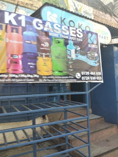 A marketing banner for "K1 GASSES" and "KOKO UPISHI MART" showcases various gas cylinders, emphasizing they offer "MORE THAN JUST GAS." The banner includes the KOKO brand logo and two Kenyan phone numbers: 0720 484 658 and 0724 630 053. The banner is displayed on a blue metal rack structure, positioned outside a building with a blue exterior and steps leading up to it.