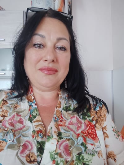 The media file contains a close-up, front-facing selfie of a woman in Sochi, Russia. She has dark hair, light brown eyes, and is wearing light-colored makeup, including eyeshadow and lipstick. She is smiling slightly and appears relaxed. She is wearing a white, button-up shirt with a floral print featuring pink, red, blue, and brown flowers. A faint scar is visible on her neck. She is also wearing silver chain earrings and has glasses perched on top of her head. The background is slightly out of focus, showing what appear to be kitchen cabinets and a white wall. The lighting suggests it is daytime.