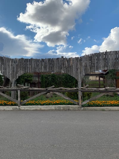 The image shows a rustic wooden fence with irregularly shaped vertical planks forming a curved barrier. In front of this fence is a lower wooden railing supported by diagonal braces, behind which a row of bright orange flowers is planted along a concrete curb. Beyond the fence, there are dense green bushes and trees, and in the distance, some buildings and structures can be seen. The sky above is blue with scattered white and grey clouds. The scene appears to be outdoors, possibly in a park or zoo setting, given the enclosure-like appearance of the wooden fence and the presence of some fenced areas behind it. The location is identified as Малахавецкі сельскі Савет, Belarus.