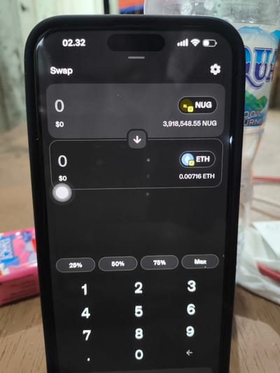 This is a close-up, eye-level, medium shot of a smartphone displaying a cryptocurrency swap interface. The phone has a black case and is held in portrait orientation. The screen shows two cryptocurrency balances: "0 NUG" with a value of "$0" and "3,918,548.55 NUG" above it, and "0 ETH" with a value of "$0" and "0.00716 ETH" below it. A down arrow icon is centered between these two balances, indicating a swap function. At the bottom of the screen, there are buttons for "25%", "50%", "75%", and "Max", followed by a numeric keypad and a backspace arrow. The phone is placed on a wooden surface, possibly a table. In the background, slightly out of focus, is a plastic water bottle with a blue and white label, and a pink and white packaged item, possibly gum or candy. The lighting appears to be natural, coming from the top. The context of Kualakeureutoe, Indonesia is not visually inferable from the image.