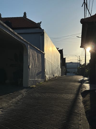 The image depicts a narrow street in Dalung, Indonesia. The street is paved with interlocking diamond-patterned pavers. Tall buildings border each side, their surfaces reflecting sunlight. Overhead power lines crisscross the sky. A building with traditional Balinese architectural elements is visible on the left. The sun is positioned at the end of the street, creating a bright glare.