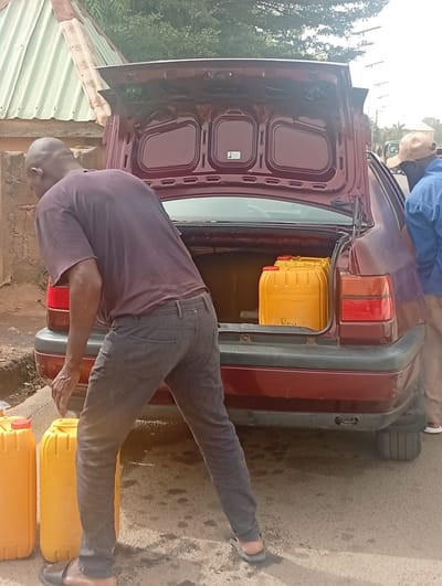 The image depicts a man unloading yellow jerrycans from the open trunk of a maroon sedan in what appears to be a roadside setting. Another man wearing a blue shirt and tan hat stands near the rear of the car. Two yellow jerrycans are already on the ground. The location appears to be a residential street, potentially in Barakin Akawo, Nigeria.