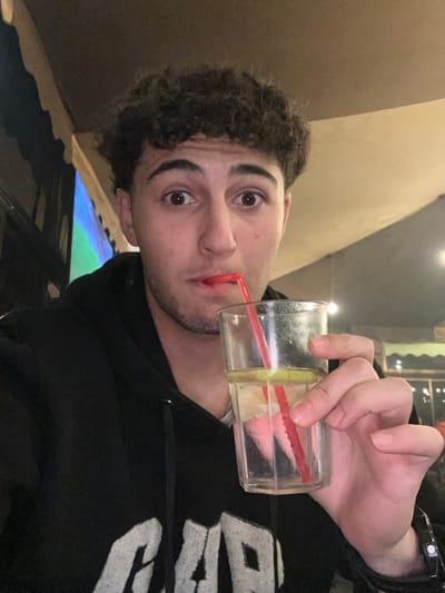 A young man with curly brown hair takes a sip from a clear glass using a red straw. The glass also contains a lemon wedge. He wears a black hoodie with white lettering. A black watch is visible on his wrist. The background is dimly lit, suggesting an indoor setting. The image is slightly tilted, with the man's face and the glass prominently featured.