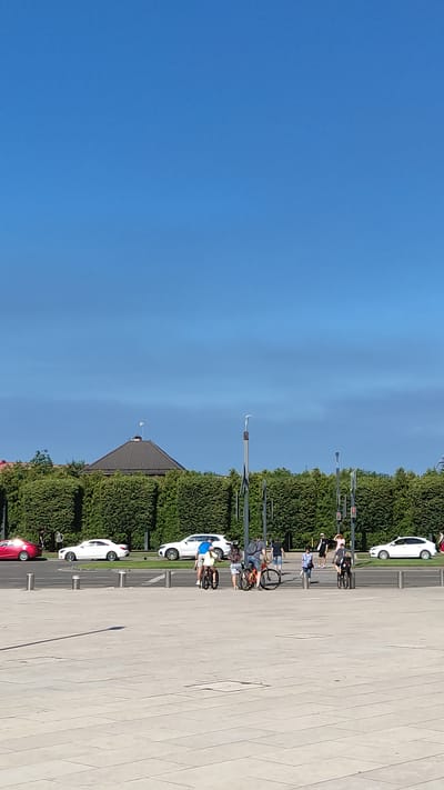 In Krasnodar, Russia, on a sunny day, a wide, paved public plaza features a line of parked cars and a crosswalk on a street bordered by a dense, manicured hedge. Several people are visible, some on bicycles and others walking, near the crosswalk and the street. A white sedan is parked behind the hedge, and a red car is visible further to the left, also behind the hedge. A brown-roofed building with a pitched roof is partially visible behind the hedge. The sky is clear and blue.