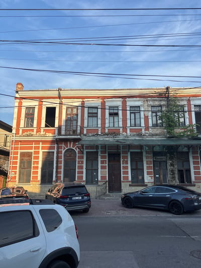 The image depicts a street scene in Chișinău, Moldova. The central subject is a weathered, multi-story building with orange and white detailing. The building exhibits signs of age, with peeling paint, dark wooden doors, and grimy windows, some of which are barred. A tree grows out of the building's facade on an upper level, adding to the impression of decay.
In front of the building, several cars are parked along the roadside. A dark-colored sedan is parked closest to the building, followed by a dark blue SUV, and then the rear of a white SUV. The cars suggest a modern presence contrasted against the old architecture.
The scene is illuminated by what appears to be natural sunlight, casting shadows and highlighting the textures of the building and pavement. The sky above is a clear blue with wisps of white clouds. The presence of numerous overhead power lines crisscrossing the frame further defines the urban setting.
Visible text includes license plates on the cars. The blue SUV's license plate reads "DXX 627". The number "24" is visible on a small placard attached to the building's exterior, possibly indicating an address or building number. The overall mood is one of urban normalcy, with a hint of historical charm and disrepair. The time of day appears to be late afternoon or early evening, given the angle of the light.