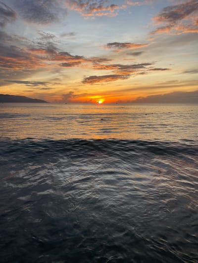 This is a vertical, full-frame photograph of a sunset over the ocean in Da Nang, Vietnam. The sun, a bright orange orb, is setting on the horizon, casting a warm glow across the sky and water. The sky is filled with clouds painted in hues of orange, pink, and yellow, with hints of blue at the top. The ocean is calm, with gentle waves reflecting the colors of the sunset. In the distance, a silhouetted mountain range forms the backdrop. A few buoys or markers are visible in the water, marking a boundary or pathway. The foreground of the image is dominated by dark, rippling water, creating a stark contrast with the vibrant sky. The scene evokes a sense of tranquility and natural beauty.