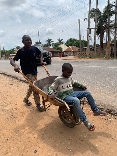 In Akwanga, Nigeria, two boys are on a road next to a dirt shoulder. One boy is sitting in an old, rusty wheelbarrow, wearing a "LUCKY" sweater, blue jeans, and Adidas sandals. The other boy is pulling the wheelbarrow, wearing a dark t-shirt with yellow stripes, patterned pants, and Adidas sandals. A black SUV is parked nearby, with power lines and palm trees visible in the background. The sky is cloudy.