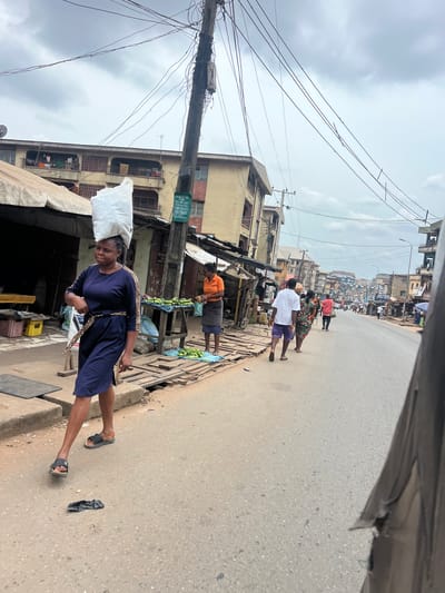 This media file captures a street scene in Nkpor, Nigeria. The primary subject is a woman in a dark blue dress with a leopard-print belt, carrying a large white bag balanced on her head. She is walking on a paved road, and her gaze is directed downwards. In the background, several other people are visible, some walking along the street and others appearing to be vendors.
The setting is a bustling urban environment, likely a market street. Buildings line both sides of the road, some appearing to be shops or residences, with signs and awnings indicating commercial activity. Several stalls are set up along the sidewalk, with one displaying what appears to be produce, possibly cucumbers. Electrical wires crisscross the sky, strung between poles, a common sight in many urban areas.
The weather appears overcast, with a muted sky suggesting it could be morning or late afternoon. The overall atmosphere suggests a typical day of commerce and movement in the area. A green sign with white text is visible on one of the buildings, but the text is largely illegible due to the image resolution and distance.
The image is captured from a slightly elevated perspective, possibly from a vehicle, as indicated by the partially visible dark frame at the top of the image. The rotation of the image suggests it was taken with a phone camera and then not reoriented.
