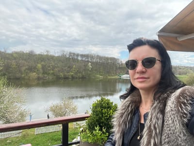 A woman wearing sunglasses and a fur-collared jacket is taking a selfie. She is on a balcony with a wooden railing. Behind her is a lake, with trees lining the opposite shore. Two figures are visible on the shoreline near the left edge of the frame, one is wearing a light blue outfit and the other is wearing black, and appear to be fishing. A white picket fence is visible below the railing. A potted plant is on the balcony. The sky is cloudy.

Without additional information, I cannot provide the exact location in Smederevska Palanka Municipality.
