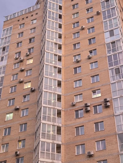 The image shows the facade of a building in Perm, Russia, which is a light-brown brick high-rise with many windows and individual air conditioning units. The building appears to be vertically mirrored at its midpoint. The windows are mostly square and aligned in rows. Some windows have curtains or blinds visible from the outside, with a couple of the windows illuminated with internal lighting. The overall perspective is from a lower angle looking up at the building. The sky is visible in the bottom-left corner, suggesting dusk or dawn.