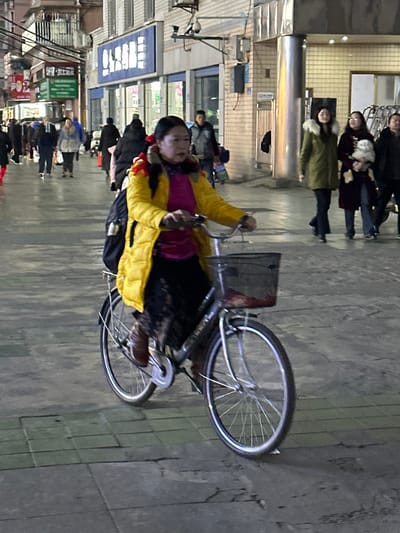 This image depicts an urban street scene at night in Jiangjin, China. The central subject is a female of apparent middle age, riding a dark gray bicycle from right to left across a paved surface. She wears a bright yellow puffer jacket over a magenta top, dark patterned trousers or a skirt, and brown shoes. Her dark hair is pulled back, secured with red accessories. A black backpack is carried on her back. The bicycle is equipped with a front basket containing a reddish object, a rear rack, and silver-spoked wheels. She holds the handlebars and appears to be pedaling.

In the background, multiple pedestrians are present. To the right of the primary subject, two females are walking; one wears a green coat with a fur-trimmed hood, and the other, in a dark coat, carries a small white, fluffy dog. Other figures are visible further in the background. The street is lined with multi-story commercial buildings featuring windows, illuminated storefronts, and signage with Chinese characters, notably a blue sign with white text on the left. Overhead utility lines are visible across the upper portion of the frame, and a security camera is mounted on a building wall in the upper right quadrant. The ground is composed of gray paving stones, illuminated by artificial light sources.