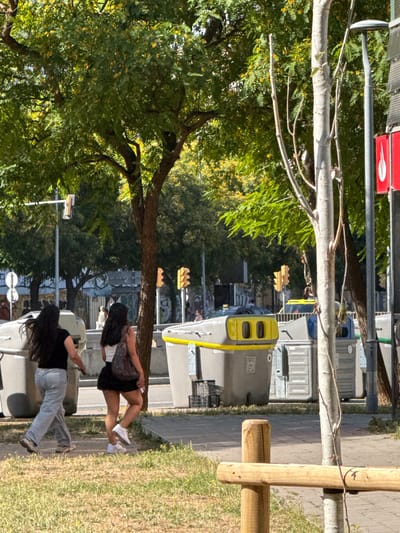 Two young women walk along a sidewalk in L'Hospitalet de Llobregat, Spain. The woman on the left has long dark hair and wears a black t-shirt and light grey sweatpants. She is walking away from the camera, and her left hand is visible. The woman on the right has long dark hair and wears a white top, a black skirt, and white sneakers. She carries a dark purse over her right shoulder and is walking away from the camera. They are walking past several large, gray and yellow public recycling bins. In the background, traffic lights are visible, along with trees and buildings. The scene is set on a sunny day, with dappled light filtering through the trees. The foreground shows a patch of dry grass and a wooden fence post.