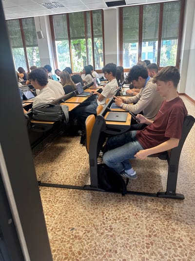 The image captures a classroom or lecture hall from a slightly elevated perspective, looking down a row of students. Numerous young adults, primarily male, are seated at individual desks with attached chairs, engaged in an activity involving digital devices.
The students are focused on laptops or tablets, which are open on their light wooden desks. Several Apple MacBooks are clearly visible, identifiable by their iconic logo. One student in the mid-ground, wearing a maroon t-shirt and jeans, is standing or kneeling on his chair, leaning over his desk and looking intently at a device. Another student nearby is pointing at a screen, suggesting discussion or collaboration. Backpacks are stowed beneath some of the chairs.
The setting is bright, with ample natural light entering through a wall of large windows. These windows feature exterior slatted blinds, common in Italy. Beyond the windows, green trees and parts of distant buildings are visible, indicating an outdoor urban or campus environment. A white speaker is mounted on the wall between window sections. The floor is a distinctive terrazzo tile with a speckled pattern of light brown, beige, and white. The ceiling has rectangular light fixtures. The scene depicts a daytime academic or training session in Milan, Italy. No specific legible text beyond the Apple logo is visible.