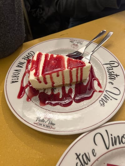 The image features a close-up, slightly angled view of a slice of cheesecake served on a white ceramic plate, indicative of an indoor dining setting. The cheesecake itself is creamy white with a visible crust and is artfully drizzled with a vibrant red berry sauce, possibly strawberry or raspberry, along with a light dusting of powdered sugar.
Two silver forks are placed on the left side of the plate, suggesting the dessert is ready to be eaten, possibly shared between two individuals. The plate's design is notable, featuring the words "Pasta e Vino" in a cursive font at the top and "COME 'NA VORTA" in block letters at the bottom. A smaller logo with "Pasta e Vino COME 'NA VORTA" is also visible. "COME 'NA VORTA" is a Roman dialect phrase meaning "like it used to be" or "like in the old days," firmly placing the scene in Roma, Italy, as indicated by the context.
The plate rests on a light brown or ochre-colored table surface. In the background, another similar plate is partially visible to the right, bearing the same "Pasta e Vino" text. To the left, out-of-focus dark fabric, possibly clothing, can be seen, suggesting a person is seated at the table. The lighting is consistent with an artificial indoor environment, likely a restaurant, creating a cozy atmosphere around the dessert. The image captures a moment of indulgence with a traditional touch.