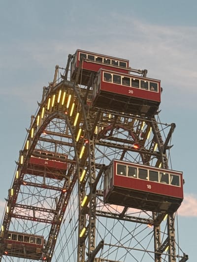 This media file features a close-up, slightly tilted view of a ferris wheel, likely the Wiener Riesenrad in Vienna, Austria. The ferris wheel has iconic red cabins, several of which are visible. One cabin has the number "18" prominently displayed, while another shows "20" vertically. The cabins have multiple windows, and faint silhouettes of people can be seen inside some of them. The ferris wheel is illuminated with yellow lights along its curved structure, suggesting it is either dusk or nighttime, or that the lights are on during the day for decorative purposes. The sky is a pale blue with wispy clouds, indicating a fair weather day. The metal structure of the ferris wheel is complex, with many supporting beams and cables. The image focuses on a section of the ferris wheel, capturing the intricate engineering and the distinctive red cabins against the backdrop of the sky.