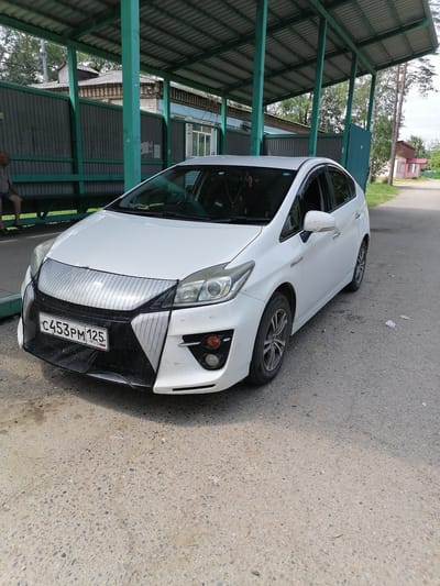 A white Toyota Prius is parked on an asphalt road in Novopokrovka, Russia. The car is positioned facing the viewer's right, with its front slightly angled towards the camera. A protective mesh cover is fitted over the car's grille and front bumper area. The car's license plate, "C453PM 125 RUS," is clearly visible on the front. To the left of the car, a covered bus stop or waiting area with green metal poles and a corrugated roof provides shade. A person is seated on a bench within this covered area. In the background, a weathered fence and a building with a light blue facade are visible. Further in the background, a small residential building with a red roof stands against a backdrop of trees and a cloudy sky. The ground is a rough asphalt surface, with some scattered debris.