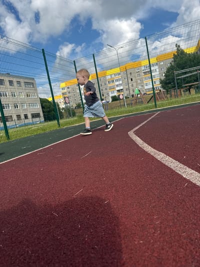 This image depicts a young child, likely a toddler, standing on a red-surfaced outdoor sports court. The child is wearing a dark grey t-shirt and light blue denim shorts. Their legs are positioned as if they are about to run or are in mid-stride, with black and white sneakers on their feet. The court has a white line marking a boundary, and a strip of green surface is visible to the left of the red court.
Behind the child, a metal fence with diamond-shaped mesh separates the court from a grassy area and residential buildings. The buildings appear to be multi-story apartment complexes with grey facades and yellow accents. The sky is visible above, with scattered white clouds against a bright blue backdrop. Sunlight indicates it is daytime.
The scene appears to be a playground or a sports area within a residential complex in Markovskii, Russia. The overall atmosphere seems casual and possibly playful, given the presence of a child on a sports court. No specific event is clearly identifiable, but the child's posture suggests movement or activity. The image is taken from a slightly angled perspective, with the child appearing to be the primary subject in the foreground.