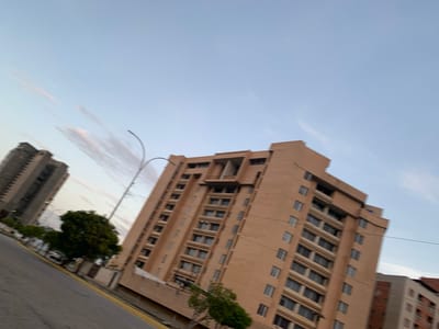 The image shows a street scene in Porlamar, Venezuela, with several tall buildings and a street in the foreground. The buildings appear to be residential or office towers, with multiple stories and many windows. The largest building in the center of the frame is a light brown brick structure with a stepped facade and balconies. To the left, another taller, gray building is visible. The sky is a light blue with some wispy clouds, suggesting it is either early morning or late afternoon. A street with a yellow curb runs diagonally across the lower part of the image. There is a tree with dark green foliage near the base of the brick building. The scene is devoid of any visible people or vehicles. The overall impression is one of an urban environment during a quiet time of day.