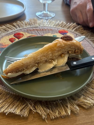 A close-up, eye-level shot captures a dessert serving on a wooden table in Heroica Puebla de Zaragoza, Mexico. The primary subject is a piece of golden-brown, glazed baklava, garnished with a single pecan nut, positioned on a round, matte olive-green plate. Beneath the baklava are several scoops of light-colored ice cream. A three-tine silver dinner fork rests on the left side of the plate. A stainless steel utility knife, marked "IKEA" on its blade and featuring a black handle with two visible silver rivets, is diagonally inserted into the baklava and ice cream. The olive-green plate sits on a circular placemat with a golden, shaggy fringe and a printed central design depicting hot air balloons, with the word "CAPPADOCIA" partially visible. In the upper left quadrant, a light-colored shallow ceramic plate is out of focus, and a clear stemmed glass is partially visible in the upper central area. A human hand, with a light skin tone and wearing a garment with visible red and grey patterns on its sleeve, is partially visible in the upper right quadrant, resting on the table.