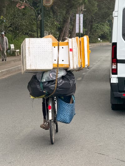 A person is riding a bicycle laden with a large amount of cargo on a paved road in Da Lat, Vietnam. The cargo includes several white rectangular boxes stacked on top of a large black trash bag and other plastic-wrapped items. A conical hat is visible among the stacked boxes. The person is wearing dark pants and brown shoes. A blue woven basket is attached to the rear of the bicycle. To the right of the bicycle, a white van is parked. The background shows trees and a sidewalk with a person walking in the distance, suggesting an urban or semi-urban environment.
