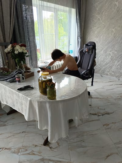 A sparsely-dressed person is seated at a large, oval-shaped table covered with a white tablecloth in a room in Andreyevo, Russia. The person, wearing glasses, is bent over, looking at a mobile phone resting on the table. A gaming chair is positioned behind the person. On the table, there is a jar of pickles, a vase with dead flowers, a mug with Cyrillic text, a TV remote, and other assorted items. The room has marble-patterned walls and marble-patterned flooring. A window is visible in the background. Curtains are to the left.