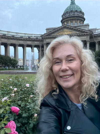A candid selfie is captured in front of the Kazan Cathedral in St. Petersburg, Russia, on an overcast day. The woman in the foreground has blonde, curly hair and is wearing a black leather jacket over a light-colored shirt. She is smiling broadly, her eyes looking at the camera. In the middle ground, a large, ornate fountain sprays water upwards, surrounded by a circular pool. Behind the fountain, the impressive facade of the Kazan Cathedral features a grand colonnade and a prominent green dome. The foreground also shows a bush with pink roses, some in bloom. People can be seen in the background, dispersed around the fountain and the cathedral's grounds.
