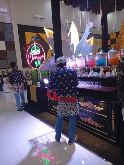 A well-lit event space features people in similar patterned shirts and jeans. A man wearing a traditional headdress stands near a beverage station with several glass dispensers filled with colorful drinks. Behind the station, there's an ornate black cabinet with gold accents and a large, stylized leaf-like decoration. A neon sign displaying a name or logo in pink and green is visible in the background, with its reflection projected on the floor.