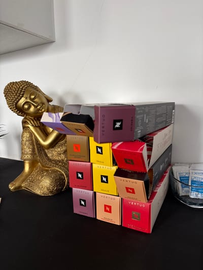 This indoor scene, located in the city of Cholula, Mexico, displays a golden Buddha statue on a black surface, positioned to the left. The Buddha is depicted reclining with its head resting on its right arm, eyes closed, and adorned with ornate golden robes. To the right of the statue, a vertical stack of Nespresso Vertuo coffee capsule boxes is arranged, featuring various colors such as purple, yellow, red, orange, and grey, with several boxes partially open. Labels visible on the boxes include "Double Espresso Chiaro," "Solelio," "El Salvador," "Colombia," "Melozio Decaffeinato," and "White Chocolate & Strawberry." To the far right, a clear glass bowl contains numerous small white packets, some labeled "Member's N Azúcar Fina" (fine sugar) and others "Edulcorante" (sweetener). The background consists of a plain white wall, which shows a faint, thin vertical crack in the upper right quadrant. A light-colored shelf or ledge is visible in the top left corner of the frame.