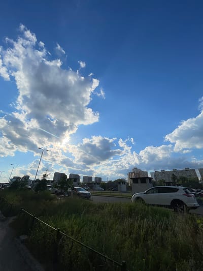 This is a vertical, low-angle photograph of a sky filled with cumulus clouds and a bright sun peeking through them. Below the sky, the scene depicts a city landscape. In the foreground, there is a patch of tall, dry grass bordering a dark metal fence. To the right of the grass is a paved area with a white SUV parked. Further back, a road is visible with several cars, including a silver sedan. Beyond the road, a row of tall, residential buildings, typical of Kyiv's urban architecture, stretches across the horizon. Streetlights are also visible. The sun's rays are creating a lens flare effect, adding a bright glow to the sky. The overall scene captures a sunny day in a residential area of Kyiv, Ukraine, possibly in the late afternoon or early evening given the angle of the sun.