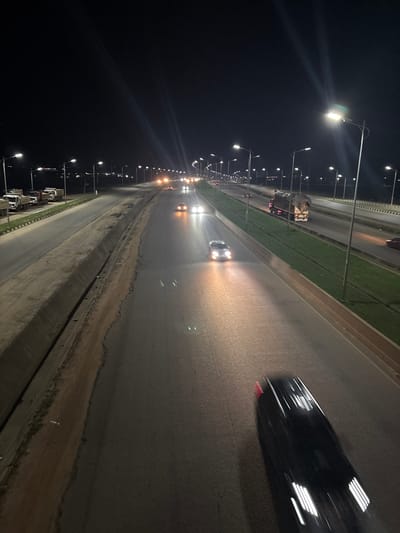 The image depicts a highway at night in Angwan Sayawa, Nigeria, with multiple lanes of traffic illuminated by streetlights. Several vehicles, including cars and trucks, are visible on the road. Some cars have their headlights on, and a few appear to be in motion, indicated by motion blur. The environment consists of a paved roadway with a grassy median and concrete barriers. Streetlights line the highway, casting light on the road and surrounding area. The scene suggests a typical night on a busy urban or suburban highway.