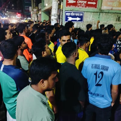 The image depicts a crowded street scene at night in Dhaka, Bangladesh. A dense gathering of men, primarily young adults, are visible in the foreground and background, suggesting a public event or gathering. The lighting is dim and artificial, with streetlights and building lights illuminating the area. Several individuals are wearing t-shirts with visible text and numbers. Specifically, one person in the foreground wears a light blue t-shirt with the text "RS RASEL SORDAR" and the number "12" on the back, along with "SPORTING CLUB" below. Other individuals are wearing brightly colored t-shirts, including yellow and orange. In the background, a blue and white sign with Bengali script and an arrow pointing to the right is affixed to a wall, indicating directions or information. The environment appears to be an urban street, with buildings and posters visible along the side of the road. The overall impression is of a lively, densely populated urban environment at night.