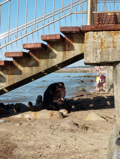 The image depicts a sunny day at a beach, likely in Zelenogradsk, Russia, given the context. In the foreground, a person in a black shirt is crouched on the sandy shore near the water's edge, appearing to be occupied with something in their hands. To the right, another person wearing a red cap and white shirt is also crouched down, seemingly interacting with the sand or water.

Behind them, a weathered concrete and rusted metal staircase ascends towards a clear blue sky. The staircase casts a shadow onto the beach. In the background, the sea stretches out, with a wooden breakwater visible in the distance. Several other people can be seen on the beach and in the water further down the coastline, suggesting a public recreational area. The overall atmosphere appears relaxed and summery. The time of day is likely midday, given the strong sunlight and distinct shadows. There is no visible text in the image.