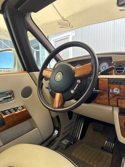 The image shows the interior of a luxury car, likely a Rolls-Royce, with a light-colored leather interior and wood trim. The steering wheel is prominent, featuring the Rolls-Royce emblem and wood accents. The dashboard includes various controls, gauges, and air vents. The car appears to be parked outdoors, possibly in a parking lot or a driveway, as indicated by the natural light and what appears to be a building or structure in the background. There are no people visible in the image. The scene is detailed and focuses on the high-end finishes of the car's interior. The mention of Dub, Montenegro, suggests a potential location for this photograph.