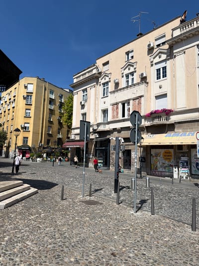 This is a sunny, outdoor, street-level view of a cobblestone plaza in Belgrade, Serbia. The plaza is flanked by buildings, with a clear blue sky overhead. 

On the left side of the image, a light yellow building stands tall. Its facade features numerous windows and balconies, and air conditioning units are visible on its exterior. In front of this building, there are steps leading up to a walkway, a lamppost, and a Western Union sign. A lone man in a white shirt and dark pants walks away from the camera on the cobblestones.

To the right of the yellow building, a cream-colored building with ornate architectural details, including classical moldings and a balcony with pink flowers, dominates the scene. The ground floor of this building houses shops with awnings. One awning is yellow and displays the word "AMANI." Various posters and graffiti adorn the storefronts. Several people are visible walking in front of these shops, including a woman in a red shirt and a person walking a small dog. A set of traffic lights are visible, and a traffic sign with two dark grey discs is placed on a pole in the plaza. 

Further into the plaza, tables and chairs are set up, suggesting an outdoor seating area for cafes or restaurants. The cobblestone pavement is the dominant ground covering. The scene appears to be a bustling urban environment on a clear day.