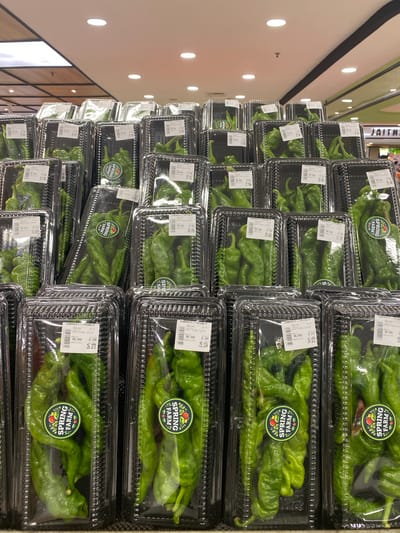 The image shows a retail display of packaged green peppers in a supermarket. The peppers are contained within clear plastic trays, which are stacked in multiple rows and columns. Each tray has a label with "SPRING FARM" branding and details about the product, including weight and price. The peppers appear to be of the shishito or a similar variety, characterized by their elongated, slightly wrinkled shape. The background suggests a well-lit commercial environment, possibly a produce section of a grocery store, with ceiling lights and shelving visible. This scene is consistent with a retail setting in North Jakarta, Indonesia, where such fresh produce displays are common.