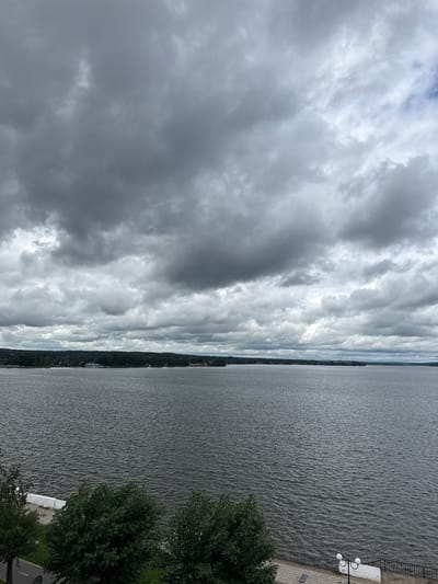 This is a landscape photograph taken from a high vantage point, overlooking a wide body of water that stretches into the distance. The sky is heavily overcast with dramatic, layered clouds in shades of gray and white, suggesting a stormy or cloudy day. The water surface is rippled, indicating a slight breeze.

Along the far bank, a thin strip of land is visible, densely covered with trees, suggesting a natural, perhaps forested, coastline. Dotted along this distant shoreline are hints of buildings, including what appear to be some taller structures that could be church spires or other urban features. Closer to the foreground, on the right side of the image, is a paved walkway and a white railing. Two lamp posts with globe lights are present on the walkway, along with a white concrete structure. A dark green tree with lush foliage is prominent in the lower right corner, partially obscuring the view of the walkway and extending towards the water.

The scene appears tranquil despite the dramatic sky, evoking a sense of quiet observation. The time of day is indeterminate due to the overcast conditions, but the light suggests daytime. There is no visible text in the image. The overall impression is one of a wide, natural expanse of water meeting a tree-lined shore under a moody sky, with a small, man-made element of a waterfront promenade in the foreground.