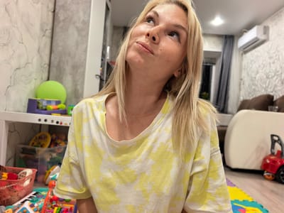 The image shows a blonde woman with light makeup, looking upwards with a thoughtful expression. She is wearing a yellow and white tie-dye t-shirt. The background suggests an indoor, possibly domestic, setting. To her left, a white shelving unit is visible, filled with various colorful toys and a large green ball. The floor is covered with a puzzle mat. In the background, a white air conditioning unit is mounted on a textured wall. A large, cream-colored sofa and a red toy car are also present. The lighting appears to be artificial, possibly from overhead lights and ambient room lighting. There is no specific indication of the city Sterlitamak, Russia, in the image itself.
