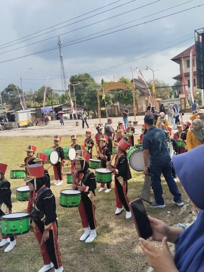 In Sambirejo, Indonesia, a youth marching band stands on a grassy field. The band members, predominantly children, wear matching uniforms consisting of black jackets with gold trim, red pants, white boots, and red and black hats. Each child carries and wears a green and white drum, holding drumsticks. An adult male stands in the middle of the band, positioned to the side of a large drum, appearing to direct the group. Other people, including adults and children, are visible in the background, observing the band. Some are taking photos or videos with their mobile phones. In the backdrop, there is a building with red and white flags. A metal archway, trees, and a utility tower are also visible in the distance. The weather appears overcast.