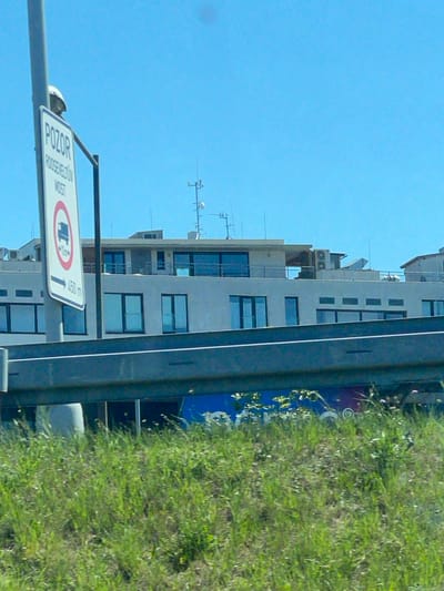 The image shows a road sign in Pilsen, Czechia. The sign reads "POZOR ROOSEVELTOV MOST" with a symbol of a truck and an arrow pointing to the left with "450 m" below it. Behind the sign is a modern building with large windows and balconies. The sky is clear and blue. In the foreground, there is green grass and a metal railing.
