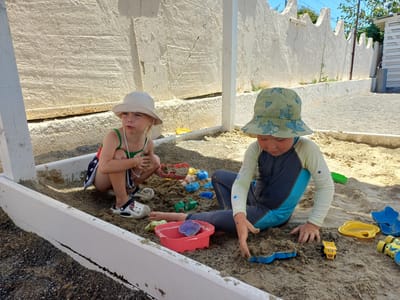 Two young children are playing in a sandpit outdoors. The child on the left, wearing a white sun hat and a green and red swimsuit, is squatting and holding a green toy. They are wearing white crocs. The child on the right, wearing a green sun hat with a shark pattern and a long-sleeved swimsuit, is sitting and digging in the sand with their hands. They are surrounded by various colorful plastic toys, including buckets, shovels, and sand molds. The sandpit is bordered by a white wooden frame, and behind it, a rough, textured white wall stretches across the background. The setting appears to be a sunny day, indicated by the bright lighting and shadows. The children's focus on their play suggests engagement and enjoyment. The location cues, such as the distinctive wall and the white beach structure, are consistent with a coastal or resort area. The time of day is likely daytime, given the strong sunlight. There is no visible text in the image.